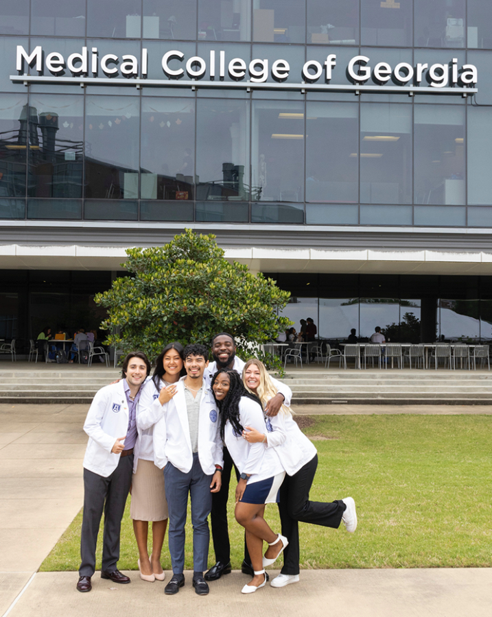 A group of six medical students strike a silly pose outside a modern-looking medical school building. They are all wearing their official lab coats.