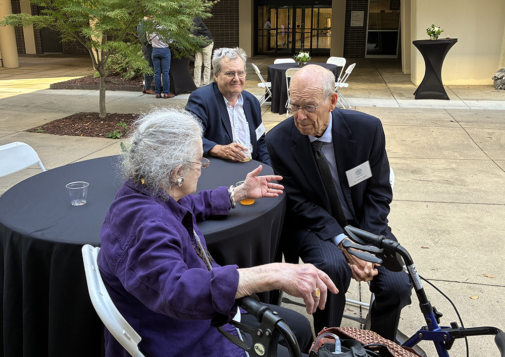 two men and woman seated at an outdoor table in conversation