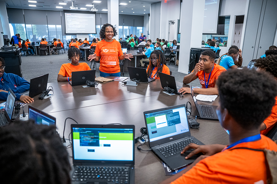 A woman speaks to a group of high school students gathered around a circular table. All the students have laptop computers and are working on completing a competition.