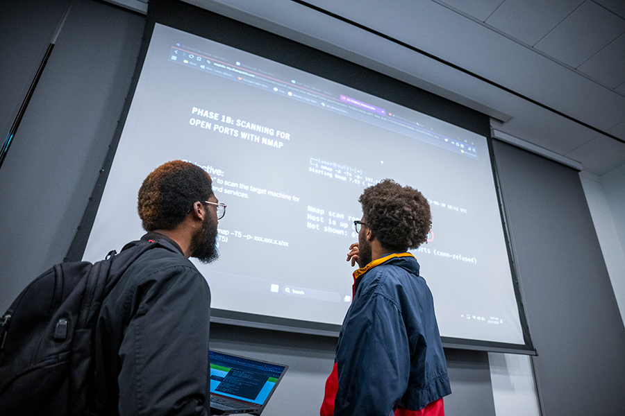 Two college-aged men stand in front of a projection screen and look at instructions for a competition they are helping with.