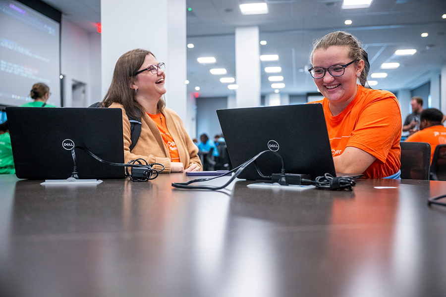 A high school teacher and a female student share a laugh during a competition. The high school girl is using a laptop computer.