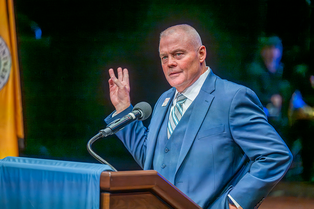 A man stands at a podium holding up three fingers.