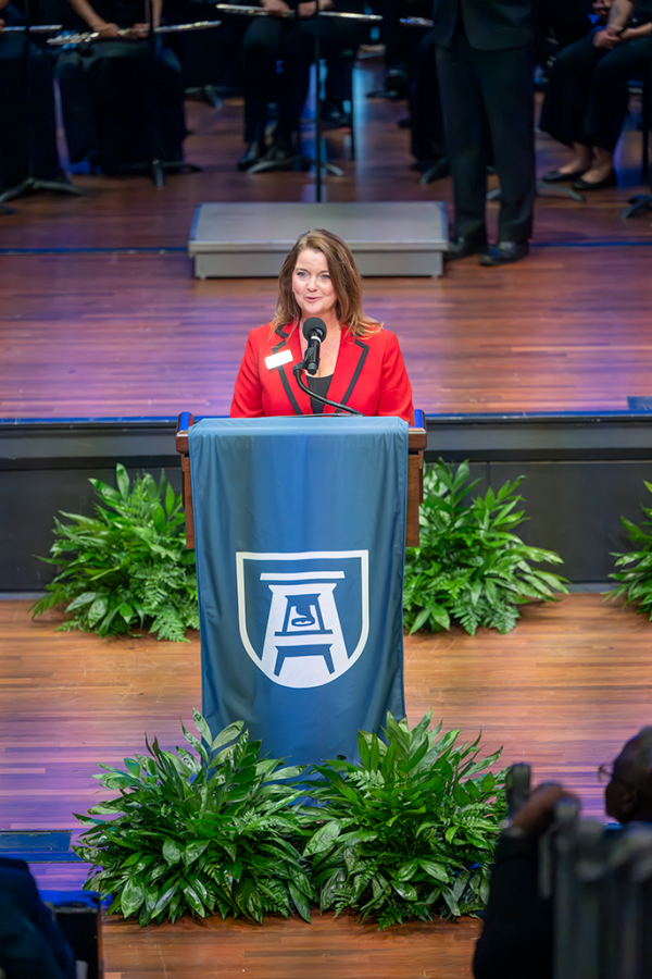 A woman stands at a podium decorated with plants, speaking into a microphone.