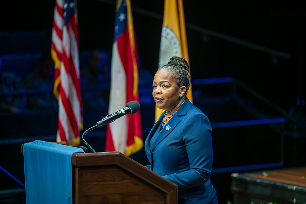 A woman stands at a podium with a microphone with flags in the background.