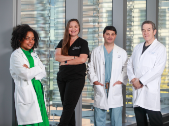 Four medical doctors, three wearing lab coats and one in medical scrubs, stand in front of a window. The group includes two men and two women.