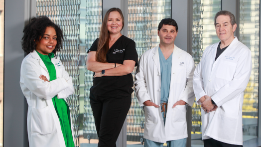 Four medical doctors, three wearing lab coats and one in medical scrubs, stand in front of a window. The group includes two men and two women.