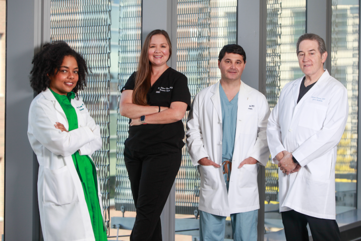 Four medical doctors, three wearing lab coats and one in medical scrubs, stand in front of a window. The group includes two men and two women.