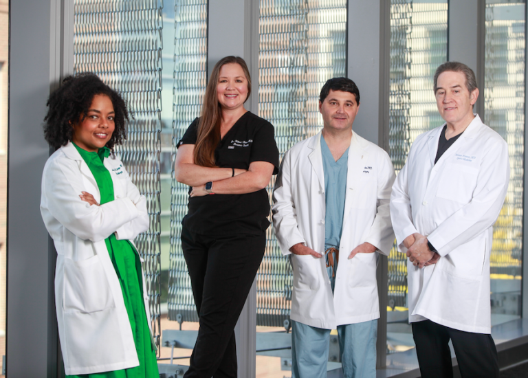 Four medical doctors, three wearing lab coats and one in medical scrubs, stand in front of a window. The group includes two men and two women.