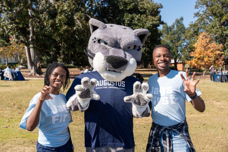 Augusta University's jaguar mascot, Augustus, and two students hold their hands like a paw.