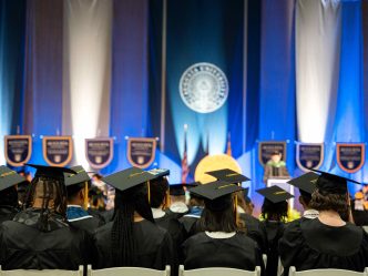Students wearing graduation caps and gowns sit at an indoor event.