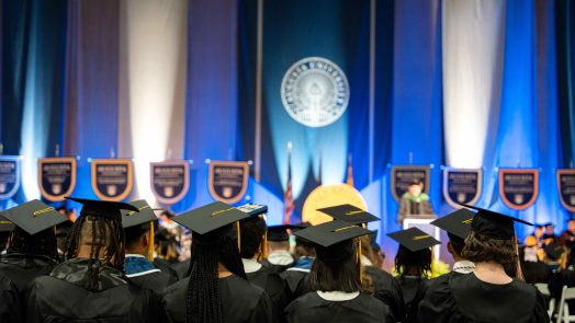 Students wearing graduation caps and gowns sit at an indoor event.