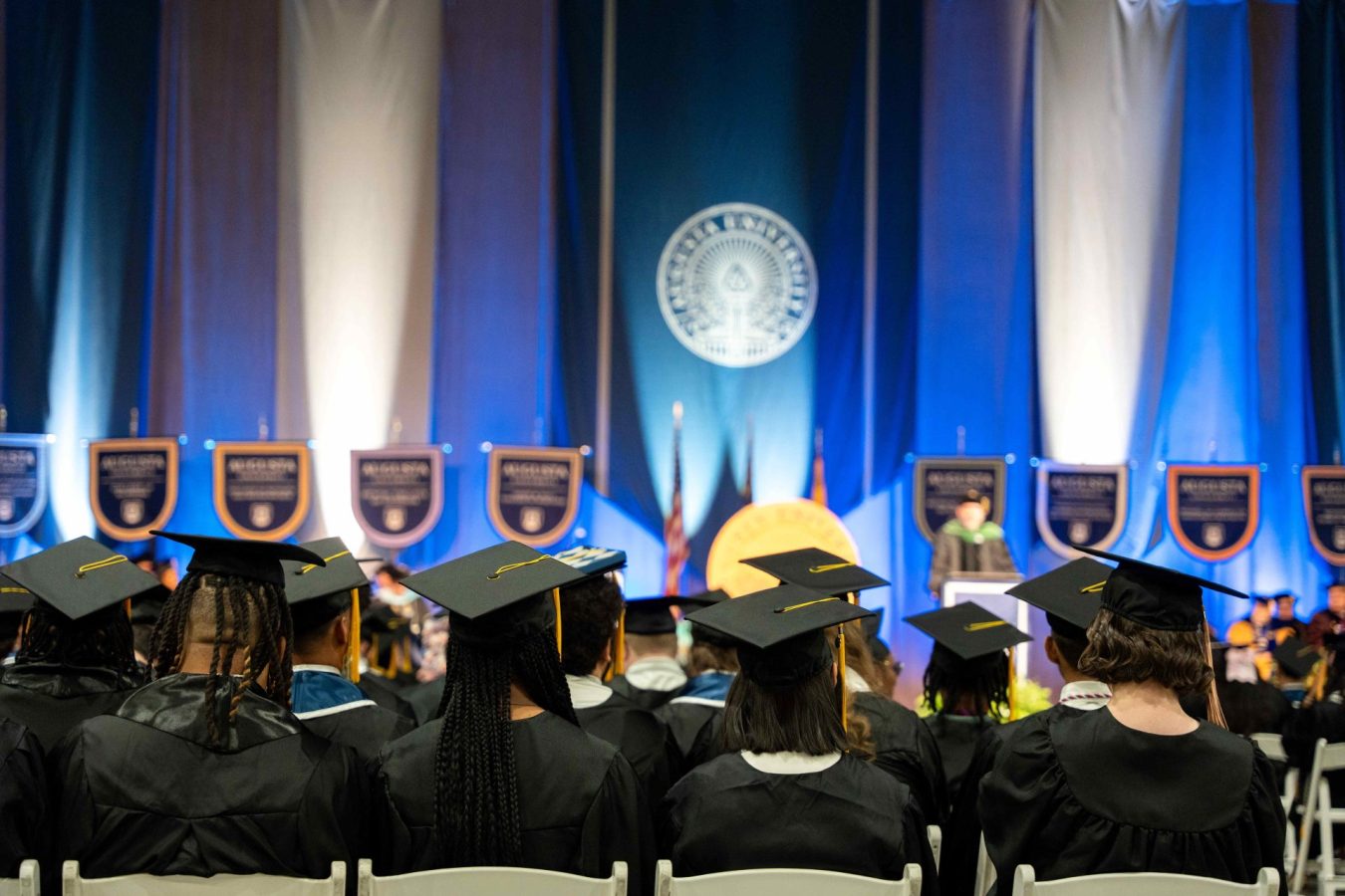 Students wearing graduation caps and gowns sit at an indoor event.