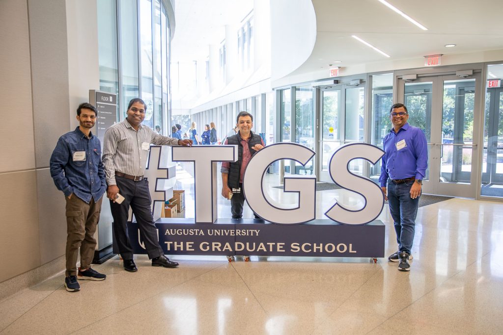 Four men stand next to a sign that reads "#TGS: Augusta University The Graduate School" in a large atrium of a modern building.