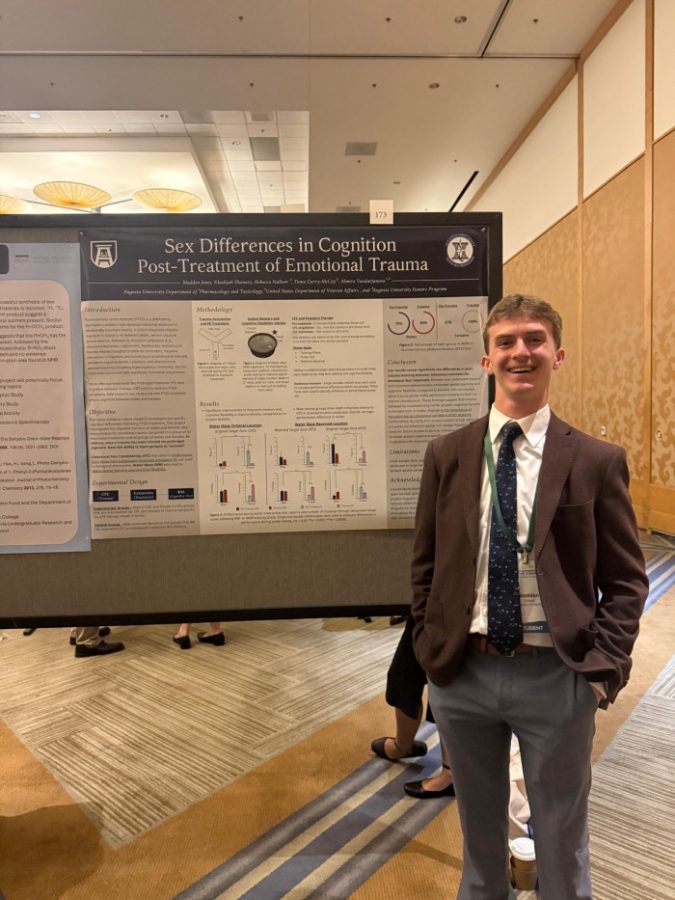A male college student stands next to a scientific poster on a stand positioned inside a large conference room.