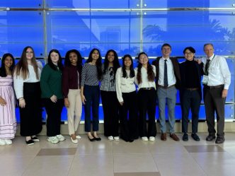 A group of eight college-aged women, two college-aged men, and a professor standing inside a long hallway at a national conference center.