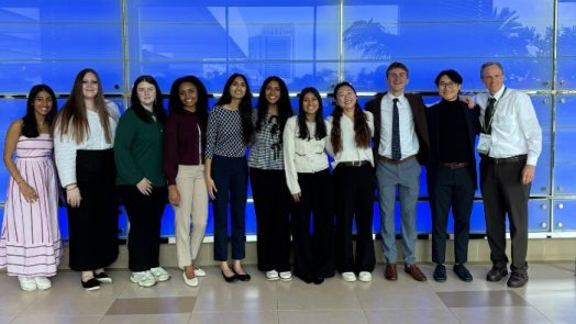 A group of eight college-aged women, two college-aged men, and a professor standing inside a long hallway at a national conference center.