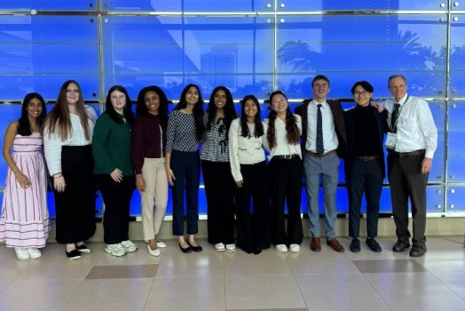A group of eight college-aged women, two college-aged men, and a professor standing inside a long hallway at a national conference center.