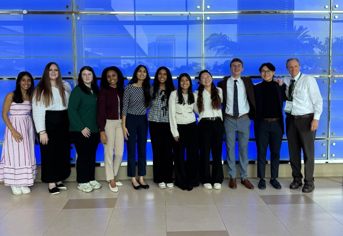 A group of eight college-aged women, two college-aged men, and a professor standing inside a long hallway at a national conference center.