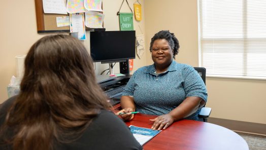 A woman smiles at a young student across from a desk.