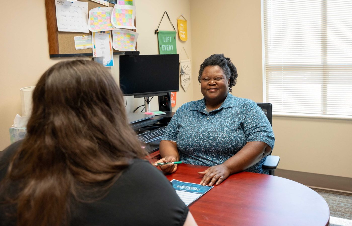 A woman smiles at a young student across from a desk.