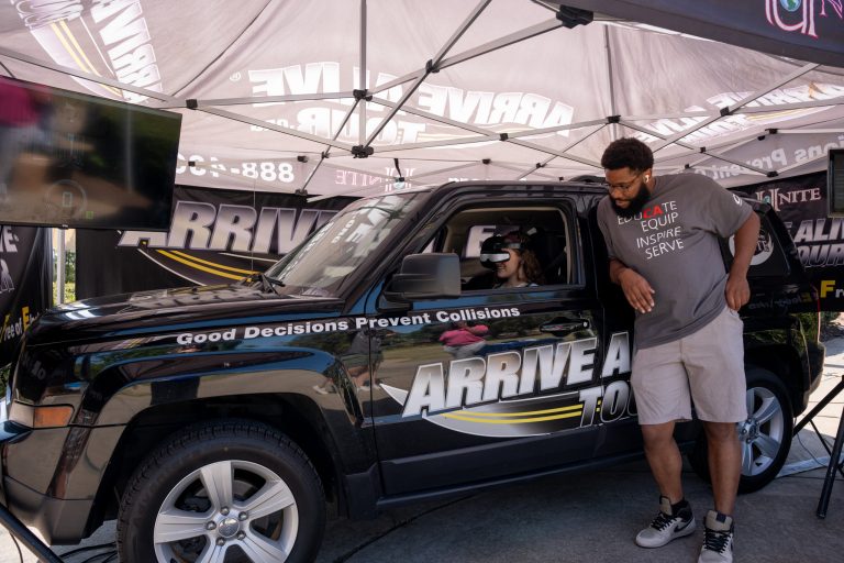 A woman wearing virtual reality goggles sits in a stationary vehicle.