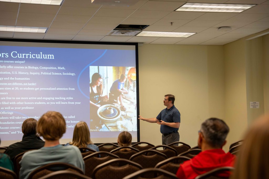 A male college staff member speaks to a large room filled with high school students and their parents on a college tour.