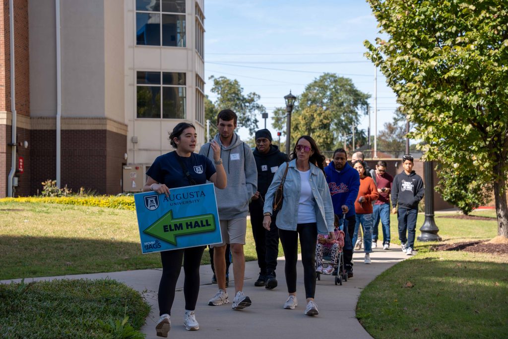 A college-aged woman leads a tour for high school students and their parents on a college campus.