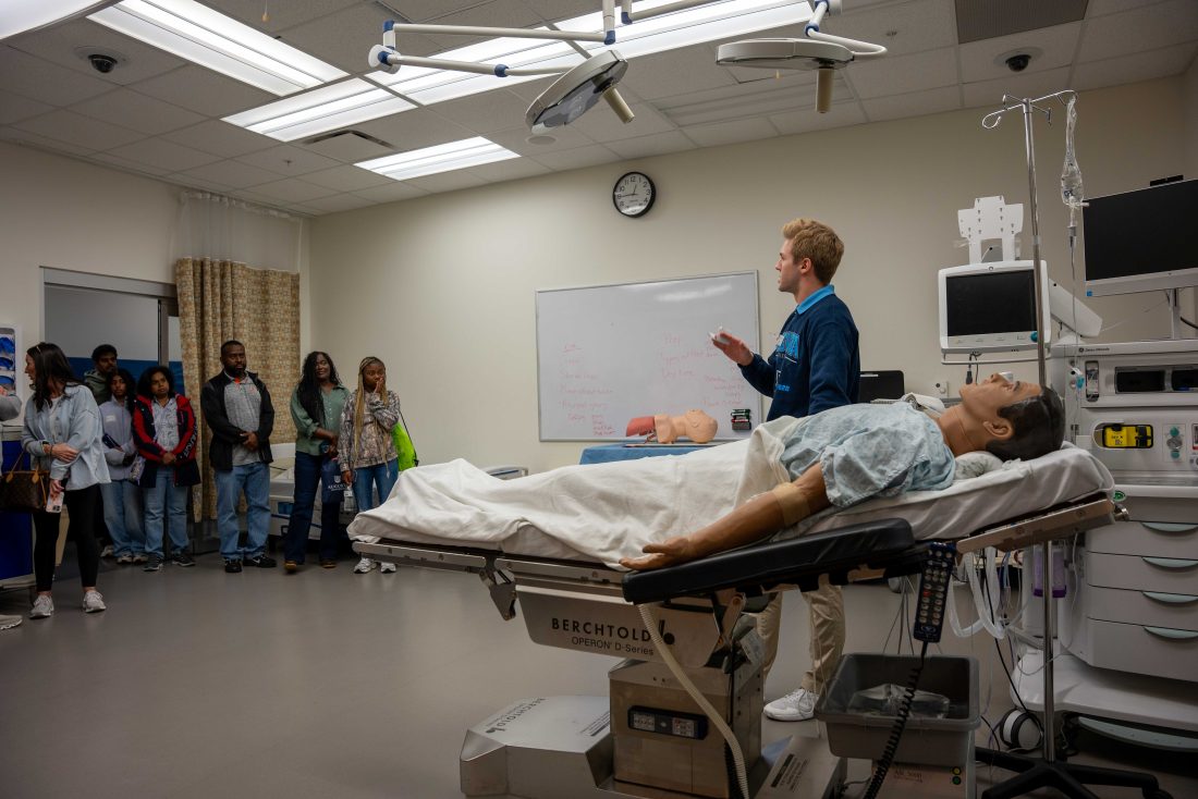 A college-aged man speaks to a great of people while standing near a medical dummy.