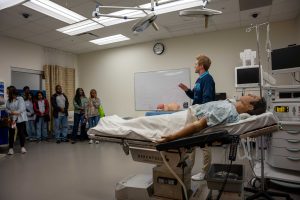 A college-aged man speaks to a great of people while standing near a medical dummy.