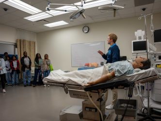 A college-aged man speaks to a great of people while standing near a medical dummy.