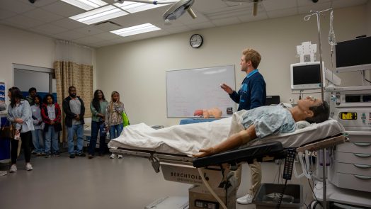 A college-aged man speaks to a great of people while standing near a medical dummy.