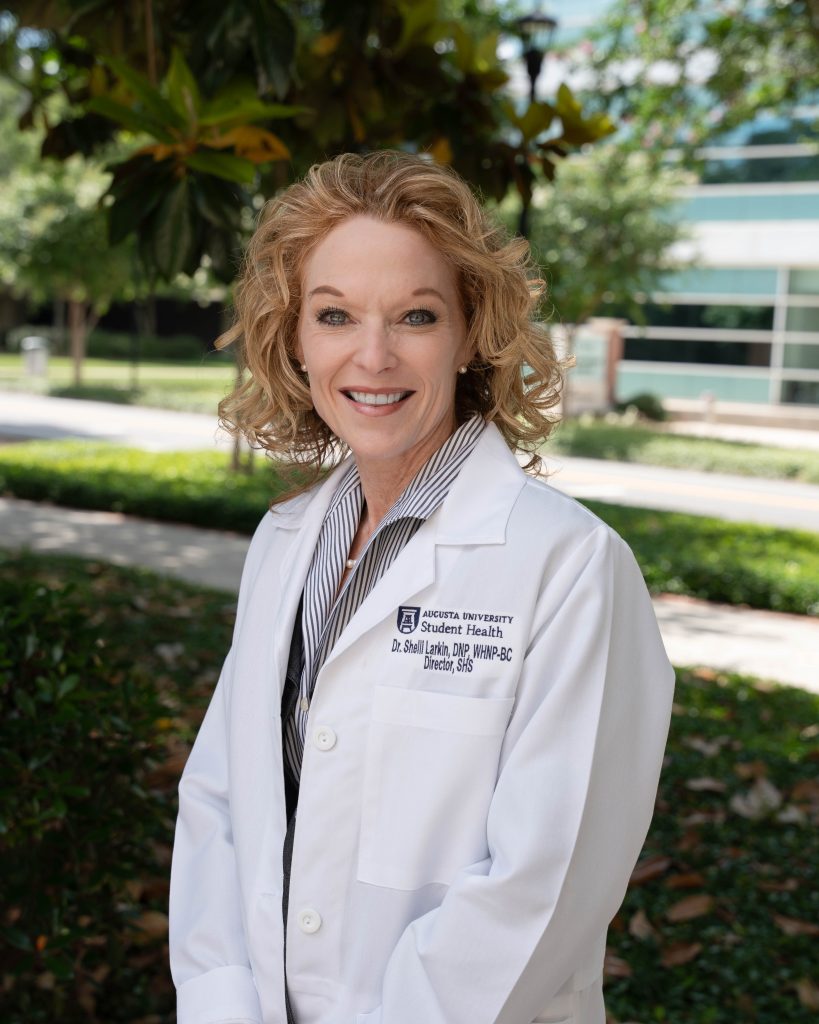 A smiling woman in a lab coat stands outside. 