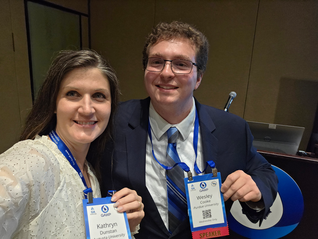A man in a suit and glasses, Wesley Cooke, and a woman in a white shirt, Katy Dunstan, holding up blue badges on lanyards to the camera at a conference.
