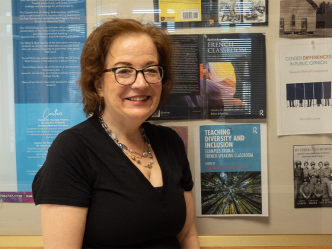 A female college professor stands in a hallway of a campus building. She is standing next to a collection of books on display, which she wrote.