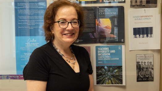 A female college professor stands in a hallway of a campus building. She is standing next to a collection of books on display, which she wrote.