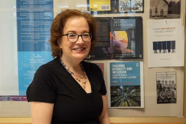 A female college professor stands in a hallway of a campus building. She is standing next to a collection of books on display, which she wrote.