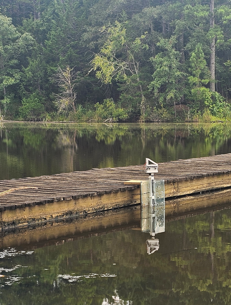 A four-foot PVC rectangular device submerged halfway into a canal, held in place by mounting it to a wooden walkway.