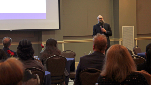 A male college professor presents data to over 100 people gathered in a large conference room.
