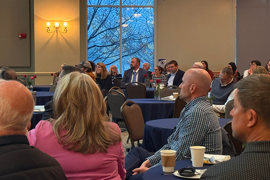 A male college professor presents data to over 100 people gathered in a large conference room.