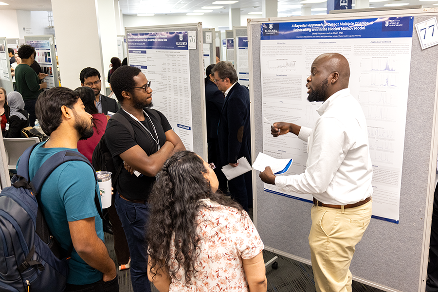 A male college student shares about his research with three fellow students during a large poster presentation.