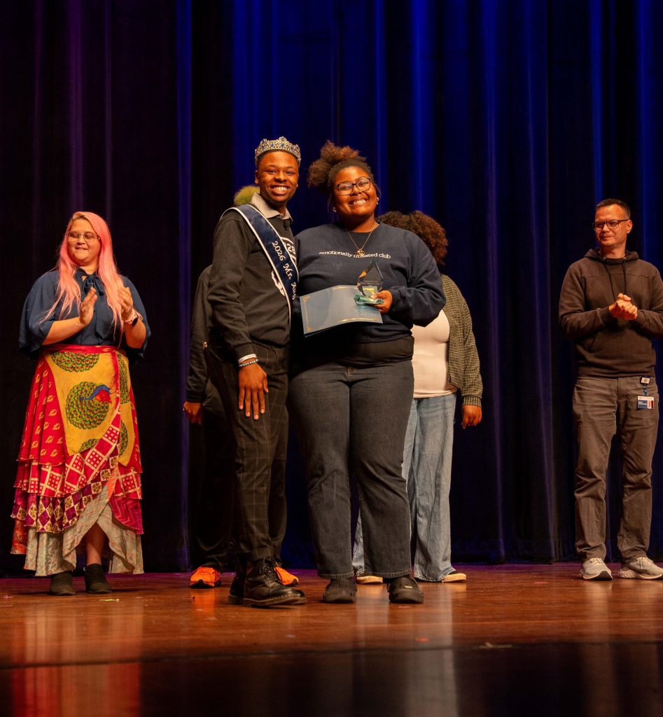 A man wearing a sash and crown smiles while standing beside a woman holding a certificate and trophy.