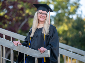 Woman stands with hands on stair railing wearing graduation cap and gown.