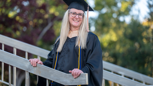 Woman stands with hands on stair railing wearing graduation cap and gown.