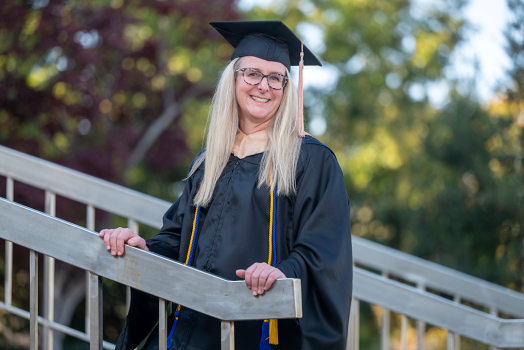 Woman stands with hands on stair railing wearing graduation cap and gown.
