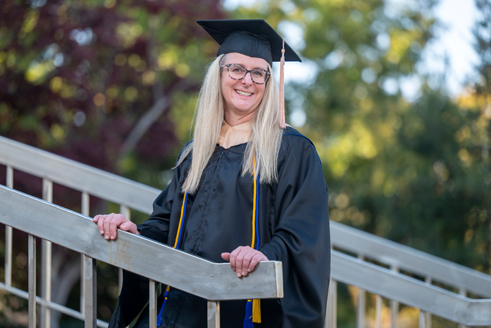 Woman stands with hands on stair railing wearing graduation cap and gown.