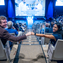 Two college students, one man and one woman, give each other a fist bump while sitting in a large auditorium. It is filled with faculty, staff and students from a university celebrating the start of a new academic year.