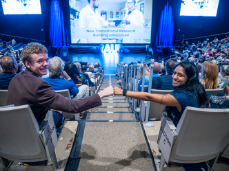 Two college students, one man and one woman, give each other a fist bump while sitting in a large auditorium. It is filled with faculty, staff and students from a university celebrating the start of a new academic year.