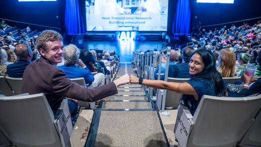 Two college students, one man and one woman, give each other a fist bump while sitting in a large auditorium. It is filled with faculty, staff and students from a university celebrating the start of a new academic year.