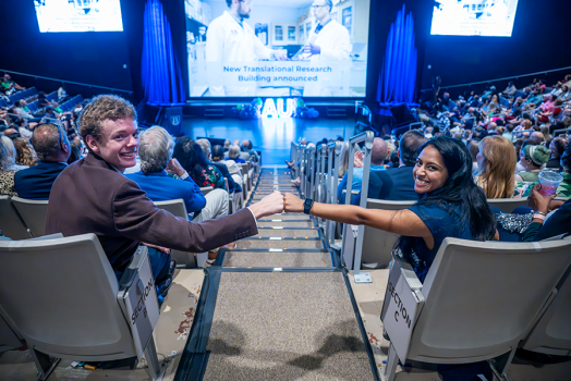 Two college students, one man and one woman, give each other a fist bump while sitting in a large auditorium. It is filled with faculty, staff and students from a university celebrating the start of a new academic year.
