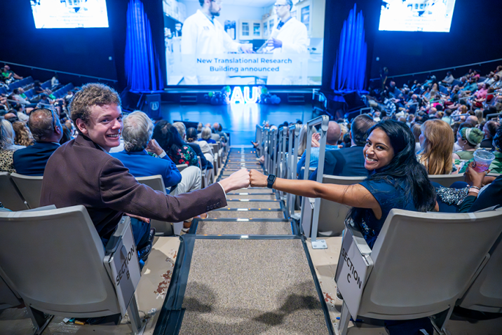 Two college students, one man and one woman, give each other a fist bump while sitting in a large auditorium. It is filled with faculty, staff and students from a university celebrating the start of a new academic year.